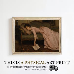 May include: A framed art print depicting a person in a long, pale pink dress sleeping on a dark green upholstered sofa. The person rests their head on a pillow, with one hand hanging down holding an open book. The frame is a metallic gold color.