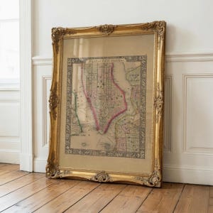Op de afbeelding: Een ingelijste vintage kaart met de titel Plan of New York, voorzien van gedetailleerde stratenplannen en gekleurde randen. De kaart zit in een sierlijke, zware goudkleurige lijst met gedetailleerde bloem- en krulmotieven langs de randen.