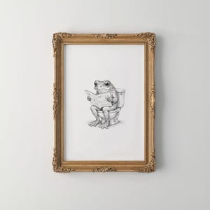 Puede incluir: Ilustraci&oacute;n en blanco y negro enmarcada que representa una rana leyendo un peri&oacute;dico en un inodoro. La obra de arte est&aacute; presentada en un marco dorado ornamentado, a&ntilde;adiendo un toque de elegancia a la escena caprichosa.