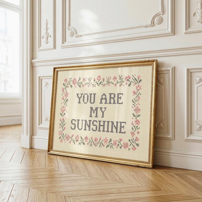 May include: A framed cross-stitch artwork with the text "YOU ARE MY SUNSHINE" in black letters. The artwork is surrounded by a floral border in pink and green, set against a cream-colored background. The frame is gold.