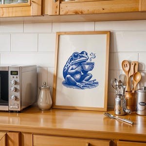 May include: A framed print of a blue frog holding a cup of coffee. The frog is sitting on a green leaf and has a steam coming from the cup. The print is on a white background and has a wooden frame.