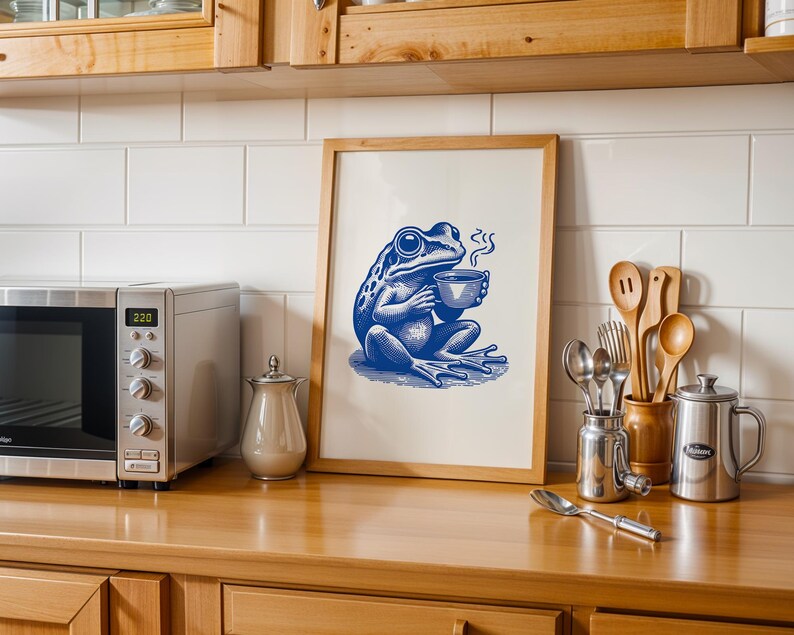 May include: A framed print of a blue frog holding a cup of coffee. The frog is sitting on a green leaf and has a steam coming from the cup. The print is on a white background and has a wooden frame.