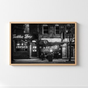 May include: Black and white framed print of a vintage building facade. The sign above the entrance reads "Cadillac Lounge" in neon. A classic car is mounted above the storefront. The image captures a retro, urban scene.