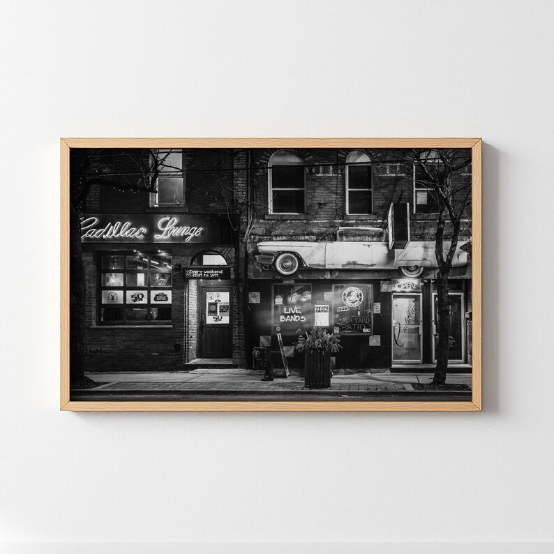 May include: Black and white framed print of a vintage building facade. The sign above the entrance reads "Cadillac Lounge" in neon. A classic car is mounted above the storefront. The image captures a retro, urban scene.