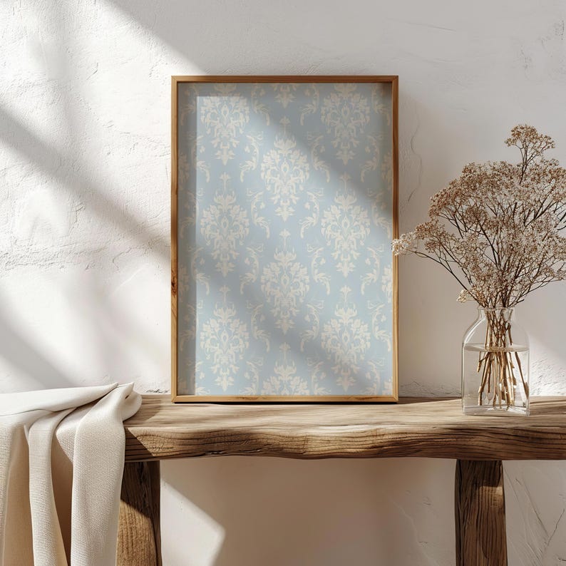 May include: A rectangular wall art print featuring a light blue background with a repeating cream-coloured damask pattern, encased in a thin wooden frame. The print displays symmetrical floral and scrollwork motifs across the entire surface.