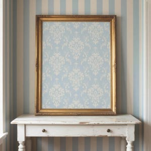 May include: A rectangular gold-toned picture frame containing a light blue and white damask patterned print. The frame features ornate, carved detailing along its edges and is positioned upright on a white wooden console table with a single drawer.