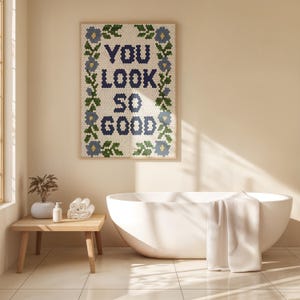 Puede incluir: Una obra de arte enmarcada con las palabras "YOU LOOK SO GOOD" en baldosas hexagonales azul marino, rodeada por un borde floral con acentos azules y verdes. La obra cuelga sobre una ba&ntilde;era blanca.