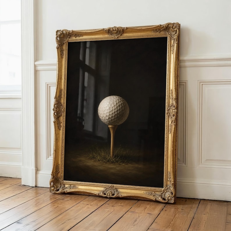 Op de afbeelding: Een ingelijst verticaal kunstwerk met een witte golfbal met putjes op een houten tee. De sc&egrave;ne is geplaatst tegen een donkere, sfeervolle achtergrond met een plukje gras aan de basis. Het geheel zit in een sierlijke, antieke gouden lijst.