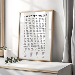 May include: A framed print titled "THE POTTY PUZZLE" features a word search puzzle with words related to potty training. The print has a light wood frame and is displayed on a white surface. The background includes a white curtain.