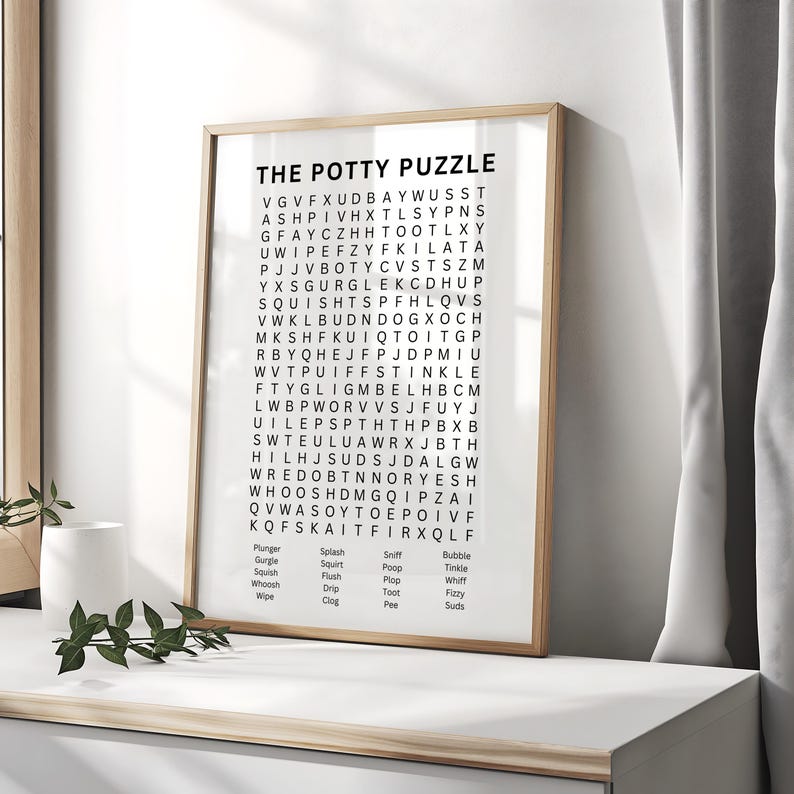 May include: A framed print titled "THE POTTY PUZZLE" features a word search puzzle with words related to potty training. The print has a light wood frame and is displayed on a white surface. The background includes a white curtain.