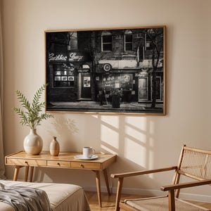 May include: A framed black and white photograph of a building with the text "Cadillac Lounge" in neon. The photograph is displayed above a wooden desk with a vase, a small vase, and a cup and saucer. A woven chair is to the right.