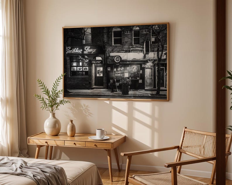 May include: A framed black and white photograph of a building with the text "Cadillac Lounge" in neon. The photograph is displayed above a wooden desk with a vase, a small vase, and a cup and saucer. A woven chair is to the right.