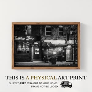 May include: Black and white art print of a building with a vintage car mounted on the side. The sign says "Cadillac Lounge" in neon. The print is framed in a wooden frame. The text at the bottom says "This is a physical art print."