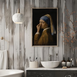 May include: A framed wall art print depicting a person wearing a blue headscarf and brown garment, holding their nose with one hand. The artwork is displayed in a bathroom setting with a white pendant light, a sink, and toiletries.
