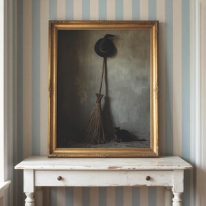May include: A framed wall art piece featuring a black pointed hat hanging above a wooden broomstick, with a small black cat sitting on the floor beside it. The artwork is enclosed in an ornate gold frame and displayed on a white wooden table.