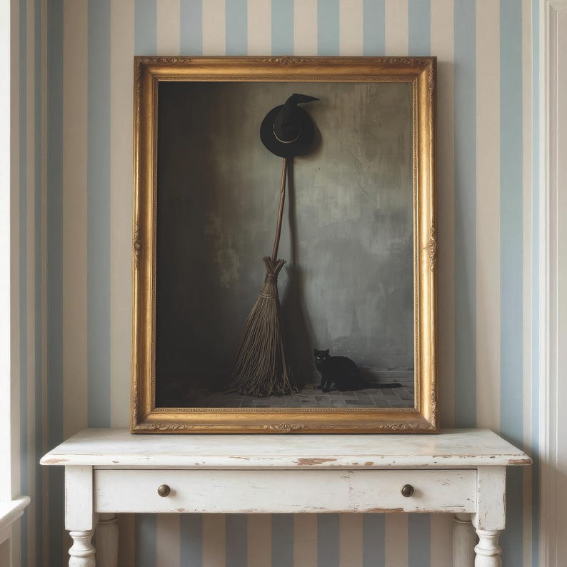 May include: A framed wall art piece featuring a black pointed hat hanging above a wooden broomstick, with a small black cat sitting on the floor beside it. The artwork is enclosed in an ornate gold frame and displayed on a white wooden table.