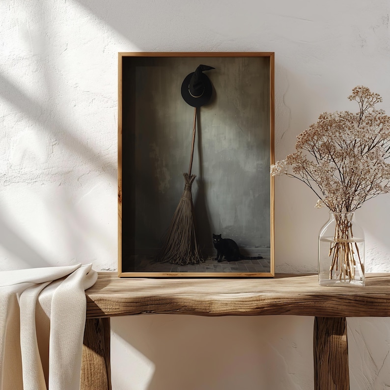 May include: A framed wall art print featuring a black witch hat hanging above a wooden broomstick, with a small black cat sitting on the floor. The artwork is displayed in a thin wooden frame, showcasing a dark, moody, and atmospheric scene.