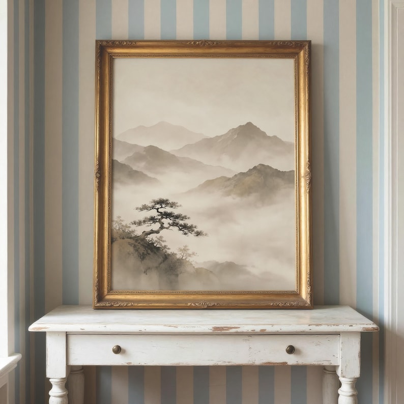 May include: A framed landscape painting in a gold-toned ornate frame featuring a monochromatic scene of misty mountain ranges and a solitary pine tree on a rocky cliffside. The artwork displays soft gray and beige tones with a hazy atmosphere.