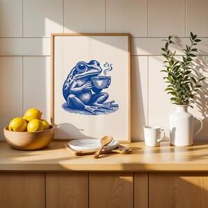 May include: A blue frog illustration holding a cup of coffee in a white frame on a wooden countertop with lemons in a bowl, a white vase with green leaves, and a white mug.