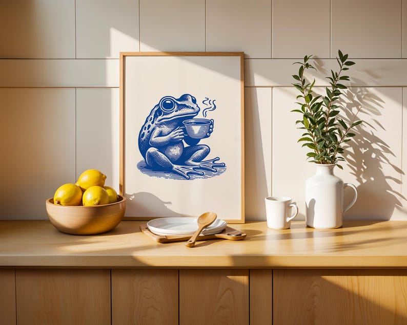 May include: A blue frog illustration holding a cup of coffee in a white frame on a wooden countertop with lemons in a bowl, a white vase with green leaves, and a white mug.