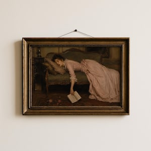 May include: A framed oil painting print depicting a person in a long pink dress reclining on a dark green sofa. The person is resting their head on a pillow while holding an open book that hangs toward the floor. The frame is ornate and metallic gold.