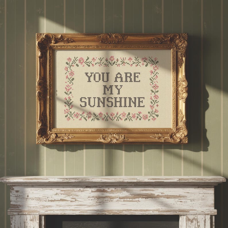 May include: A framed cross-stitch with the words "YOU ARE MY SUNSHINE" in black letters. The cross-stitch is surrounded by a floral border in pink and green. The frame is ornate and gold-colored, hanging on a green and white striped wall.