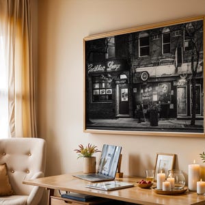 May include: A black and white framed print of a vintage storefront with the text "Cadillac Lounge" above the entrance. The print is displayed on a beige wall above a wooden desk with a laptop, candles, and other decorative items.