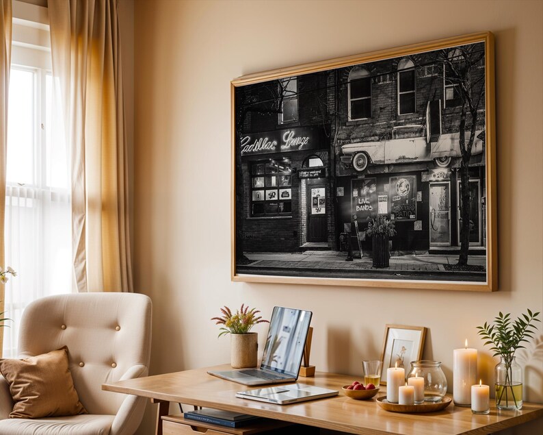 May include: A black and white framed print of a vintage storefront with the text "Cadillac Lounge" above the entrance. The print is displayed on a beige wall above a wooden desk with a laptop, candles, and other decorative items.