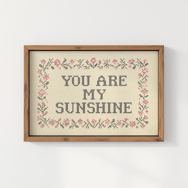 May include: A framed cross-stitch artwork with the words "YOU ARE MY SUNSHINE" in dark letters. The artwork is surrounded by a floral border in pink and green, set against a beige background.