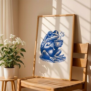 May include: A framed illustration of a blue frog holding a cup of coffee. The frog is sitting on a white background with a light blue shadow. The frame is made of wood and has a white mat.
