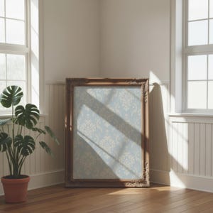 May include: A rectangular picture frame with an ornate, carved brown wooden border. The frame contains a light blue insert featuring a repeating cream-coloured damask pattern. The frame is positioned on a wooden floor in a corner of a room.