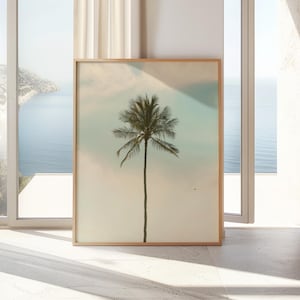 May include: A framed art print featuring a tall palm tree against a soft, pastel sky. The artwork is displayed in a light-colored wooden frame, with a view of the ocean and sky visible through a nearby window.
