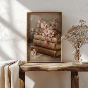 Op de afbeelding: Een ingelijste kunstdruk met een olieverfschilderij van drie gestapelde antieke boeken met verweerde kaften, bekroond met delicate lichtroze kersenbloesemtakken. De compositie bevat verspreide bloemblaadjes en een zachte, neutrale achtergrond.