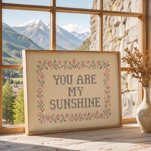 May include: A framed cross-stitch artwork with the text "YOU ARE MY SUNSHINE" in black letters. The artwork is surrounded by a floral border in pink and green. The frame is light brown, and the artwork is displayed on a wooden surface near a window.
