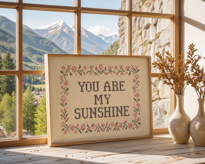 May include: A framed cross-stitch artwork with the text "YOU ARE MY SUNSHINE" in black letters. The artwork is surrounded by a floral border in pink and green. The frame is light brown, and the artwork is displayed on a wooden surface near a window.