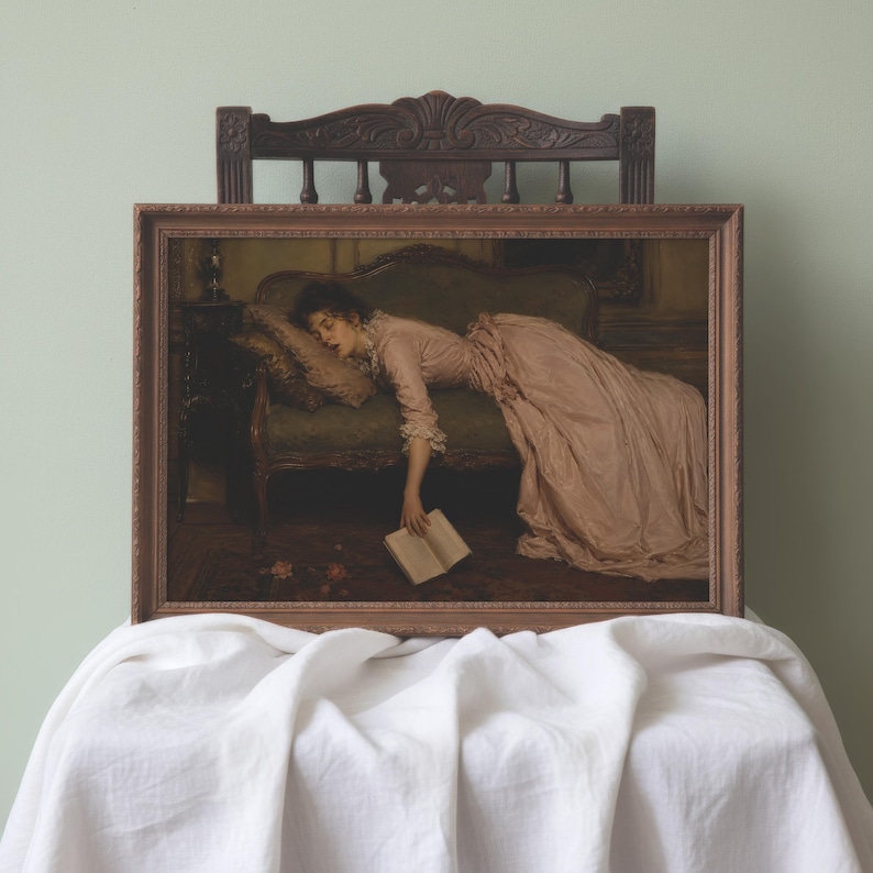 May include: A framed rectangular print featuring a painting of a person in a long, pale pink dress lying on a dark green sofa. The person is resting their head on a pillow and holding an open book that hangs toward the floor. The frame is brown.