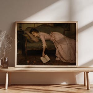 May include: A framed horizontal wall art print depicting a painting of a person in a long pink dress reclining on a green upholstered sofa. The person is asleep with one arm hanging down, holding an open book near the floor in a dimly lit room.