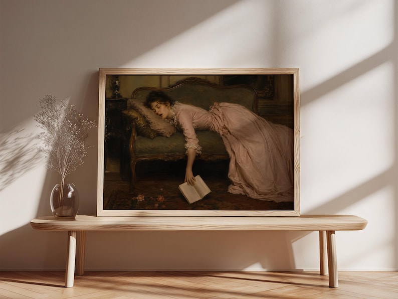 May include: A framed horizontal wall art print depicting a painting of a person in a long pink dress reclining on a green upholstered sofa. The person is asleep with one arm hanging down, holding an open book near the floor in a dimly lit room.