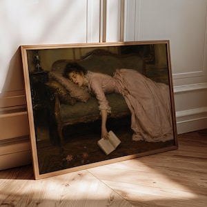 May include: A framed art print depicting a person in a long, pale pink dress reclining on a dark green upholstered settee. The person is resting their head on a pillow while holding an open book that hangs toward the floor near scattered petals.