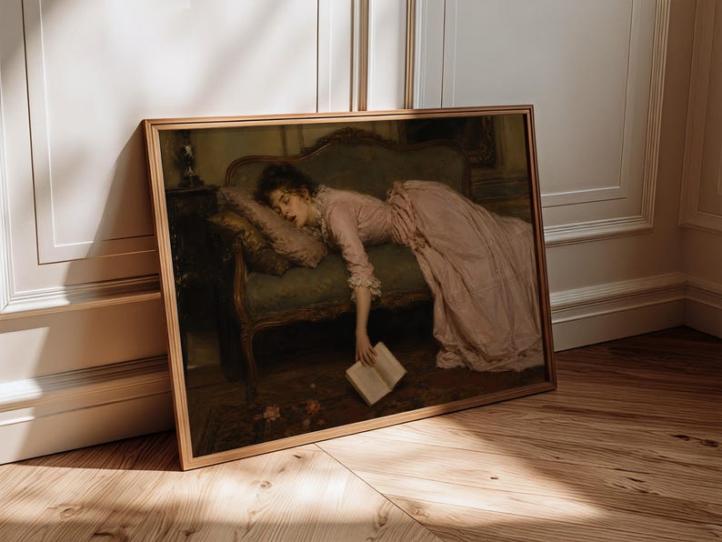 May include: A framed art print depicting a person in a long, pale pink dress reclining on a dark green upholstered settee. The person is resting their head on a pillow while holding an open book that hangs toward the floor near scattered petals.