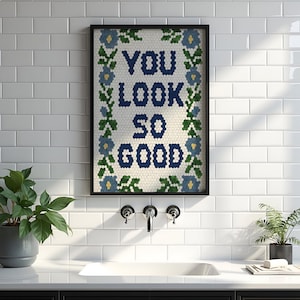 Puede incluir: Obra de arte enmarcada con el texto "YOU LOOK SO GOOD" en azul marino, rodeada por un borde floral. La obra de arte est&aacute; en un marco negro y cuelga de una pared de azulejos blancos. Una planta en maceta es visible a la izquierda.