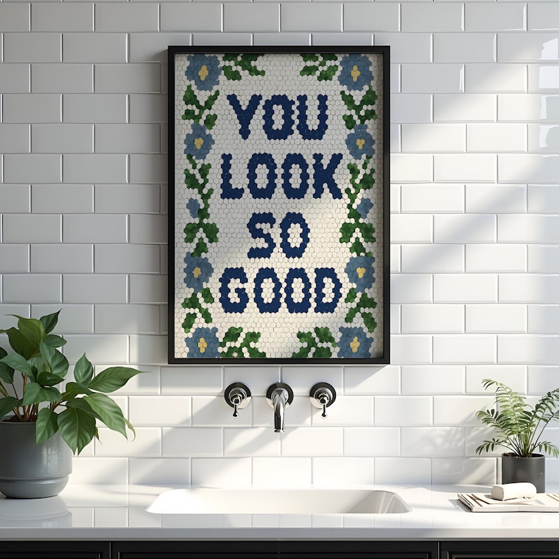 Puede incluir: Obra de arte enmarcada con el texto "YOU LOOK SO GOOD" en azul marino, rodeada por un borde floral. La obra de arte est&aacute; en un marco negro y cuelga de una pared de azulejos blancos. Una planta en maceta es visible a la izquierda.