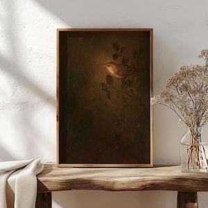 May include: A framed art print featuring a detailed illustration of a bird perched on a branch with leaves. The artwork has a warm, earthy color palette with brown and green tones. The frame is made of wood and the print is displayed on a wooden table.