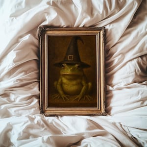 May include: A framed wall art print featuring a green frog wearing a black pointed witch hat with a square buckle. The artwork is enclosed in an ornate, metallic gold-toned rectangular frame with intricate detailing along the edges.
