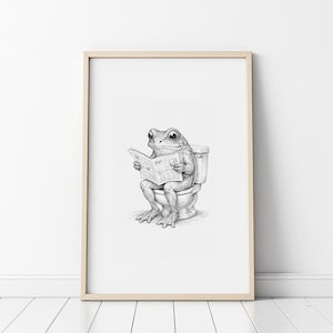 Puede incluir: Impresi&oacute;n en blanco y negro enmarcada de una rana sentada en un inodoro, leyendo un peri&oacute;dico. La rana y el inodoro est&aacute;n representados con sombreado detallado. La obra de arte est&aacute; enmarcada en un marco de madera clara.