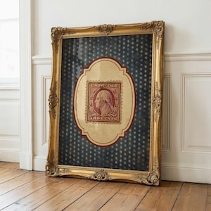 Op de afbeelding: Een ingelijste vintage Amerikaanse postzegel met George Washington, tegen een marineblauwe achtergrond met gouden sterpatronen. De postzegel zit in een decoratieve, sierlijke gouden lijst, leunend tegen een witte muur en een houten vloer.