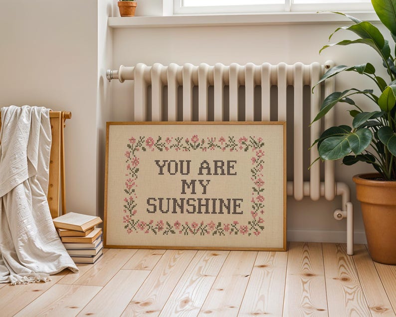 May include: A framed cross-stitch artwork with the words "YOU ARE MY SUNSHINE" in dark gray. The artwork is surrounded by a floral border in pink and green. The frame is a light brown color. The artwork is displayed on a wooden floor.