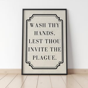 May include: Framed wall art with the text "WASH THY HANDS, LEST THOU INVITE THE PLAGUE." The text is in black, centered on a beige background, and framed in a black, decorative border. The frame is black.