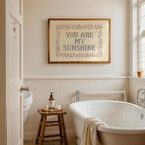 May include: A bathroom scene featuring a framed cross-stitch artwork that reads "YOU ARE MY SUNSHINE." The artwork is surrounded by a floral border. A wooden stool holds toiletries, and a white bathtub is in the foreground. The room has a neutral color palette.