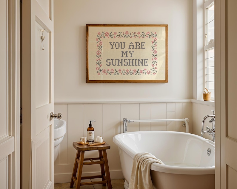 May include: A bathroom scene featuring a framed cross-stitch artwork that reads "YOU ARE MY SUNSHINE." The artwork is surrounded by a floral border. A wooden stool holds toiletries, and a white bathtub is in the foreground. The room has a neutral color palette.